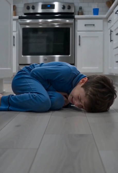 A young boy in blue pajamas is curled up on the cold kitchen floor, both hands pressed tightly against his stomach. His face is pale, twisted in pain, and his voice barely rises above a whisper as he cries out, “Dad… it hurts so bad.”