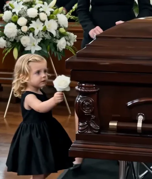 Teresa violently shoves Nina away from the sealed coffin. Nina crashes onto the carpet, her white flower falls beside her, and Teresa steps between the girl and the coffin.