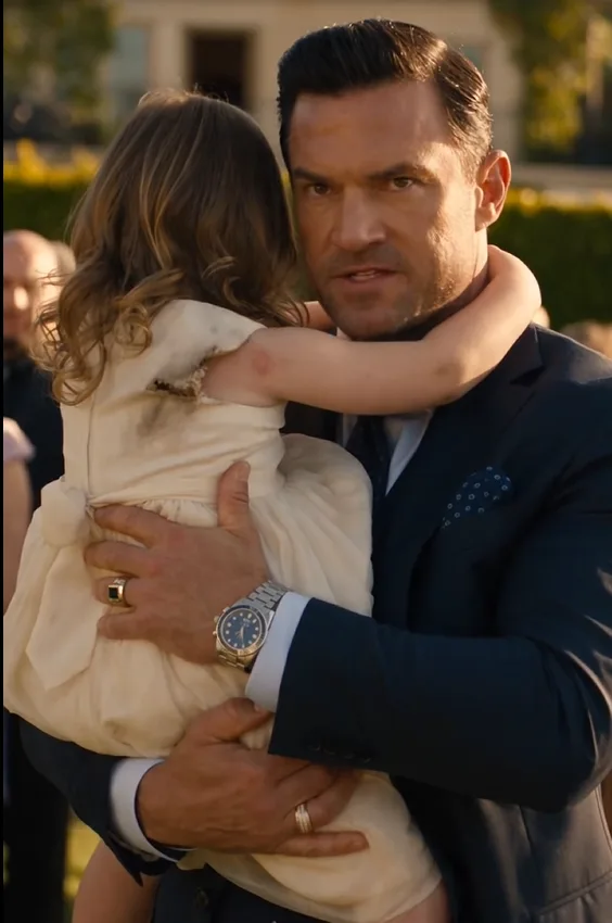 Dominic Ravello carries his three-year-old daughter into his engagement party with one shoulder of her late mother’s dress torn and one cheek visibly swollen.